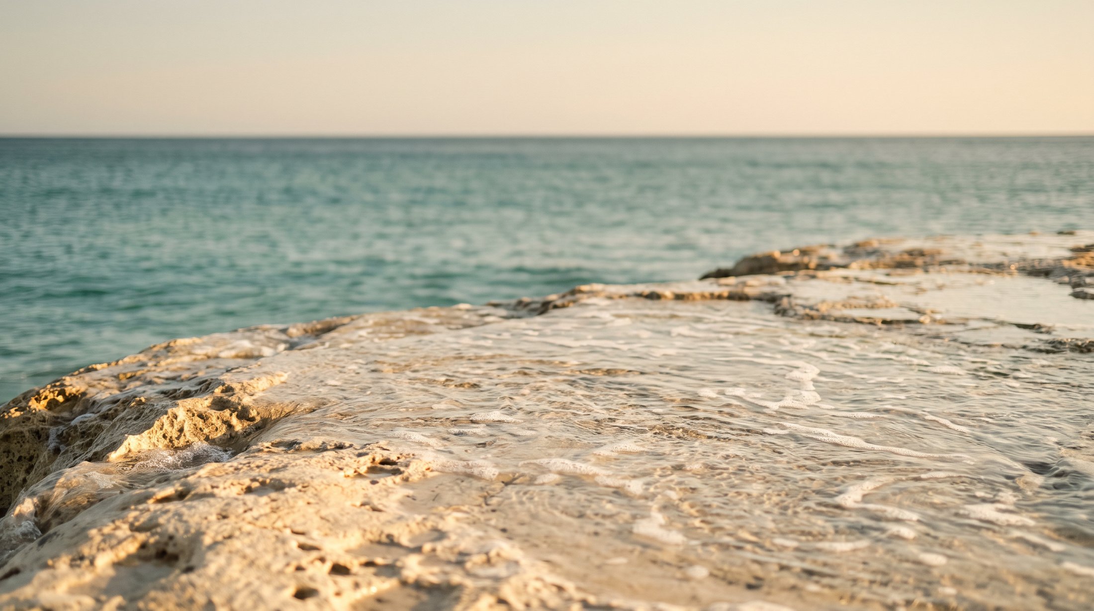 Warm travertine stone washed by clear Mediterranean water under a hazy golden horizon